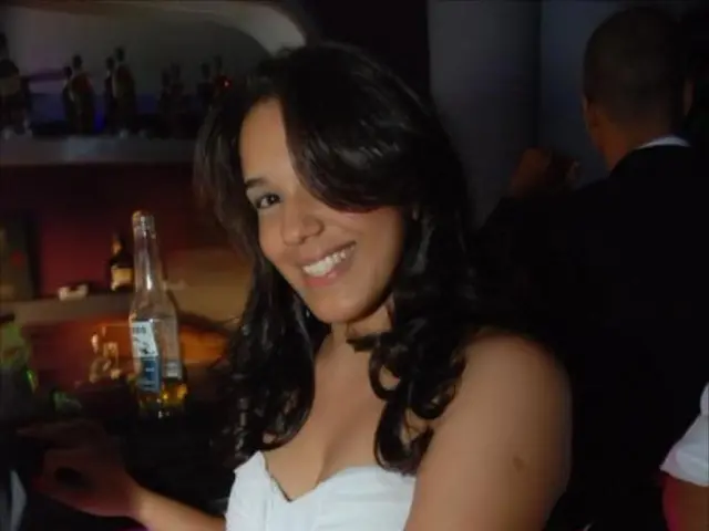 In the image we can see there is woman who is sitting on a chair and she is smiling and behind her...