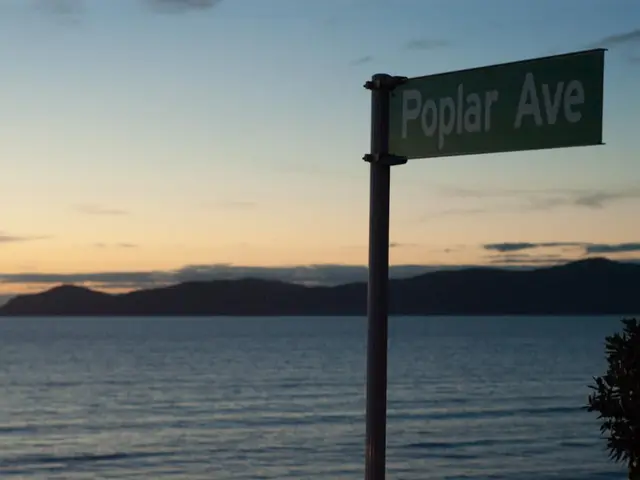 In this image we can see a sign board to the pole. Here we can see trees, water, hills and sky with...